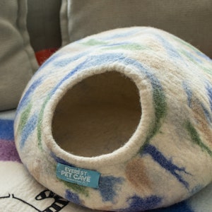 May include: A white, blue and green patterned felt cat bed with a round opening. The bed has a blue label that says "Everest Pet Cave" with the words "Handmade in Nepal" in smaller font.