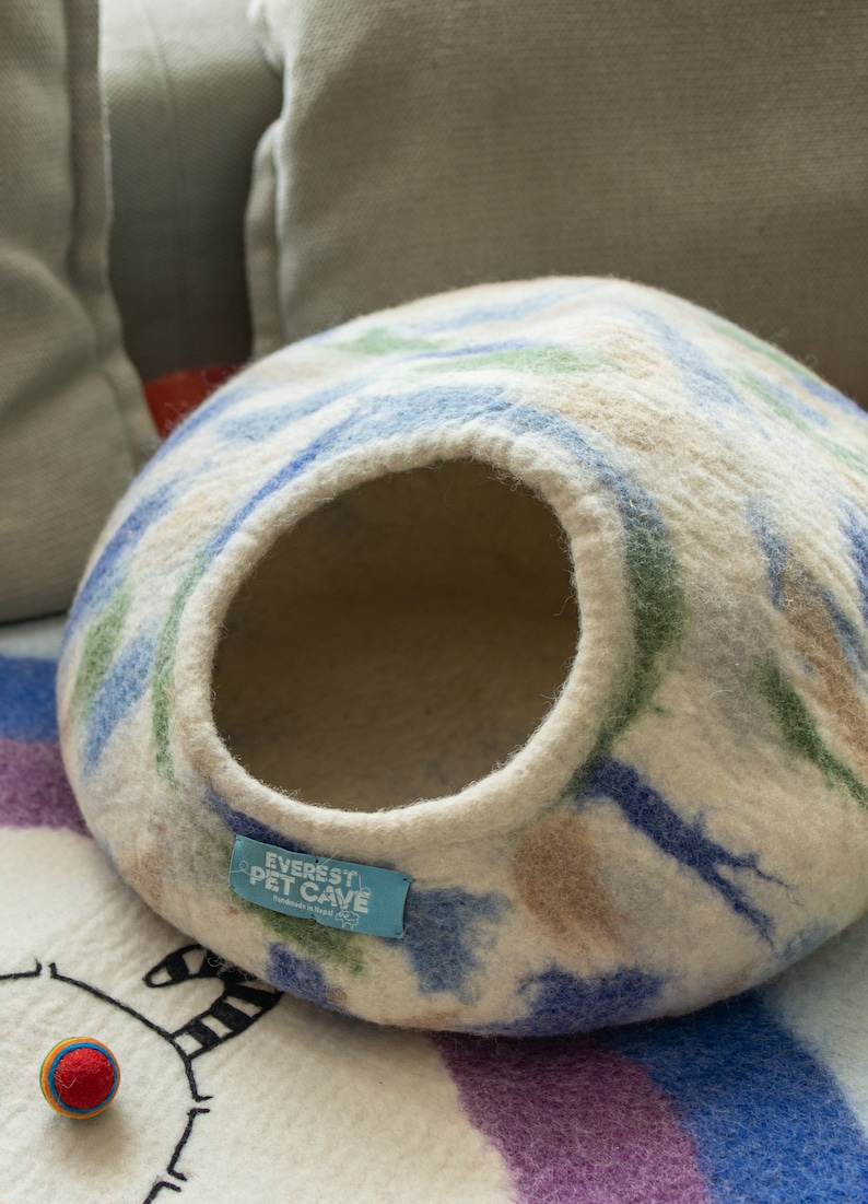 May include: A white, blue and green patterned felt cat bed with a round opening. The bed has a blue label that says "Everest Pet Cave" with the words "Handmade in Nepal" in smaller font.
