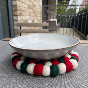 Puede incluir: Un cuenco de cerámica blanca con un diseño moteado gris se asienta sobre un posavasos redondo de fieltro de colores. El posavasos está hecho de bolas de fieltro rojo, blanco y verde.