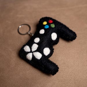 May include: A black felt keychain shaped like a video game controller with white flower accents and a silver key ring. The controller has four colored buttons in the center.