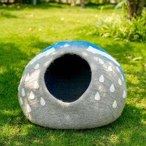 May include: A gray pet bed with a round opening, decorated with white raindrop shapes and a blue and white cloud design on top. The pet bed is made of felt and sits on green grass.