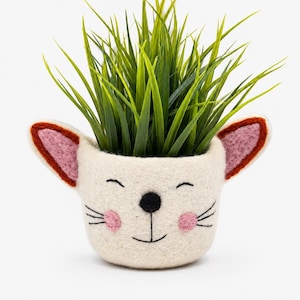 Può includere: Un'originale fioriera in feltro bianco sporco a forma di muso di coniglio sorridente, con guance rosa, baffi e orecchie con dettagli rosa e marroni. La fioriera è riempita con erba artificiale verde brillante.