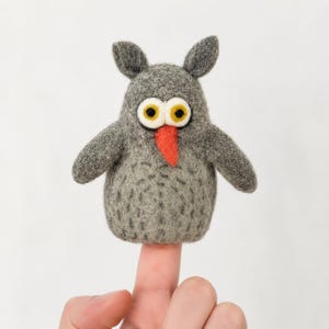 May include: A gray felt owl finger puppet with large yellow and black eyes and a bright orange beak. The owl has small, pointed ears and outstretched arms. The puppet is designed to fit on a finger.