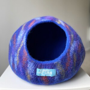 May include: A blue, purple, and orange felted cat bed with a round opening. The bed has a label that reads "Everest Pet Cave Handmade in Nepal".