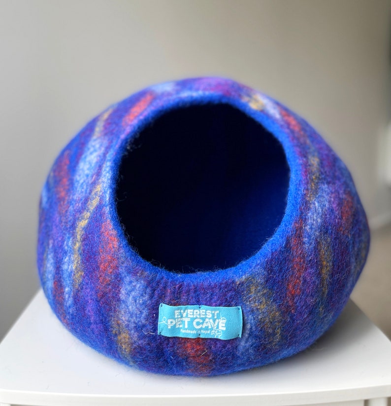 May include: A blue, purple, and orange felted cat bed with a round opening. The bed has a label that reads "Everest Pet Cave Handmade in Nepal".