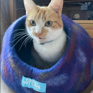 May include: A ginger and white cat with green eyes sits in a blue and purple felt cat bed. The bed has a blue tag that says "Everest Pet Cave Handmade with Love".