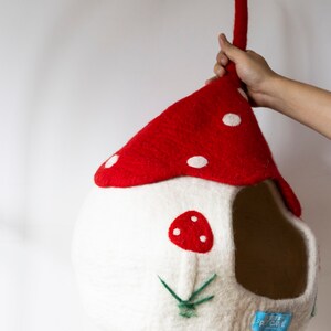 May include: A whimsical, handmade pet cave shaped like a mushroom house. The red roof has white polka dots, and the white body features a red mushroom applique and green embroidered grass. A blue tag reads "FOREST PET CAVE".