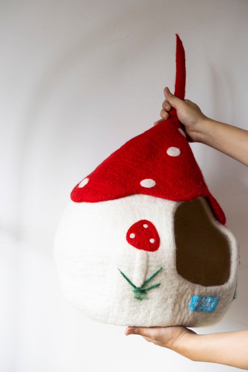 May include: A whimsical, handmade pet cave shaped like a mushroom house. The red roof has white polka dots, and the white body features a red mushroom applique and green embroidered grass. A blue tag reads "FOREST PET CAVE".