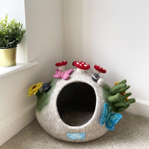以下が含まれることがあります： 緑と茶色の外側にグレーのフェルトで作られたペット用洞窟。洞窟は丸い開口部があり、フェルト製のキノコ、蝶、ひまわりで飾られています。洞窟には「エベレストペットケーブ」と書かれた青いタグが付いています。