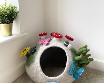 Puede incluir: Una cueva para mascotas de fieltro gris con un exterior verde y marrón. La cueva tiene una abertura redonda y está decorada con setas, mariposas y girasoles de fieltro. La cueva tiene una etiqueta azul que dice "Everest Pet Cave".