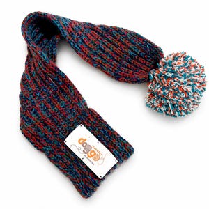May include: A knitted scarf featuring a multi-coloured yarn blend of blue, red, and green. One end of the scarf is finished with a large, round pom-pom in white, blue, and orange. A white rectangular tag is attached, displaying the text dog9 Everest accessories.