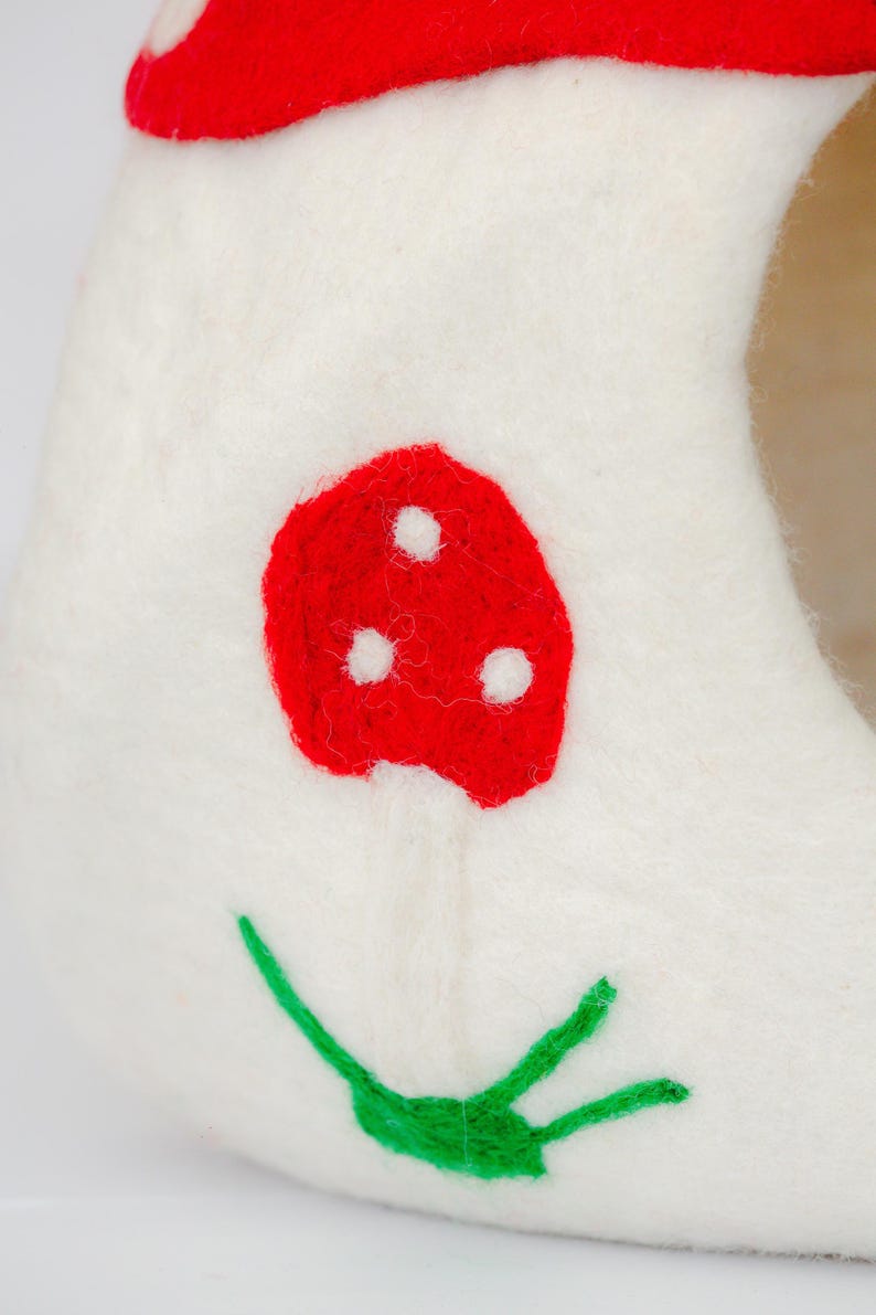 May include: A white felt mushroom-shaped cat bed with a red mushroom cap and a red mushroom on the side. The mushroom has three white dots. Green leaves are embroidered at the base. A whimsical and cozy pet accessory.