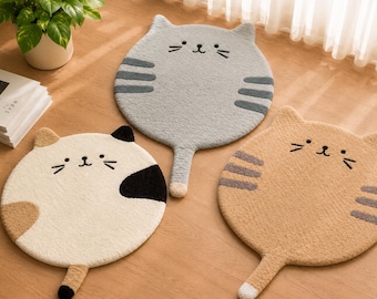 May include: Three circular rugs shaped like cat faces with ears and tails. One rug is light gray with dark gray stripes, one is cream with brown and black patches, and one is tan with gray stripes. Each rug features embroidered eyes and whiskers.