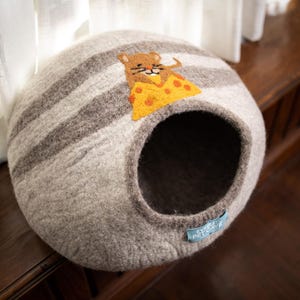 May include: A gray and white striped cat bed with a round opening. The bed has a felted texture and features an appliqué of a brown mouse holding a slice of yellow cheese. A small blue tag is attached to the bed.