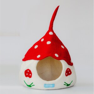May include: A whimsical pet cave shaped like a mushroom. The red top is dotted with white spots, and the base is white with a round opening. Two small mushroom appliqu&eacute;s and green leaf accents decorate the front. The cave has a tall, curved stem.