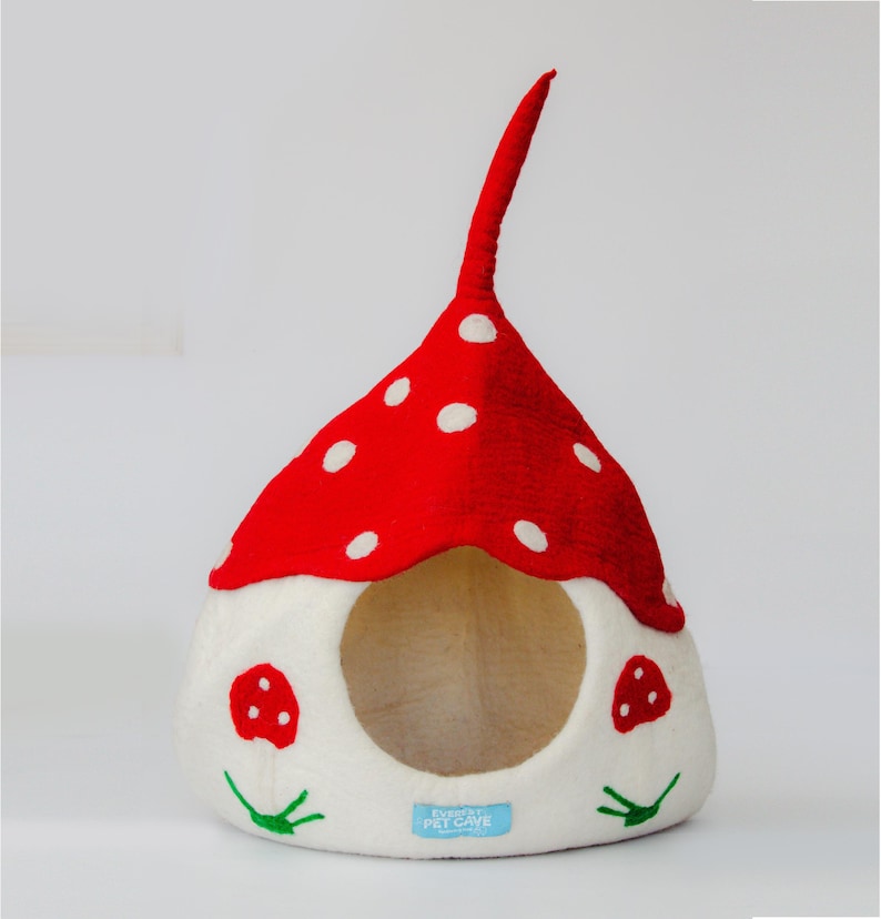 May include: A whimsical pet cave shaped like a mushroom. The red top is dotted with white spots, and the base is white with a round opening. Two small mushroom appliqu&eacute;s and green leaf accents decorate the front. The cave has a tall, curved stem.