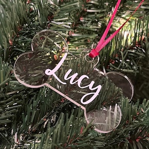 May include: Clear acrylic dog bone ornament with the name "Lucy" in white script. The ornament hangs from a red ribbon and is displayed on a green Christmas tree.