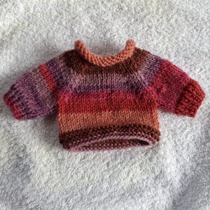 Könnte beinhalten: Ein kleiner, handgestrickter Pullover mit einem melierten Garnmuster und horizontalen Streifen in den Farben Pfirsich, Magenta, Dunkelviolett und Dunkelbraun. Das Kleidungsstück hat einen Rundhalsausschnitt, kurze Ärmel sowie gerippte Bündchen und Saum.