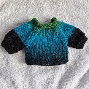 Könnte beinhalten: Ein kleiner, handgestrickter Pullover mit einem Farbverlaufsmuster, das von einem leuchtenden Grün am gerippten Halsausschnitt zu einem hellen Türkis in der Mitte übergeht und in schwarzen Rippbündchen sowie einem schwarzen Rippsaum endet.