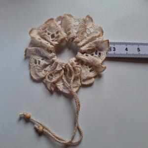 Può includere: Scrunchie in pizzo avorio con un design a balze e chiusura con coulisse. Lo scrunchie ha un diametro di circa 7,6 cm. Due piccole perline di legno sono attaccate all'estremità del cordoncino.