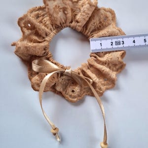Könnte beinhalten: Ein beigefarbenes, gerüschtes Haargummi aus Lochstickerei mit einer Satinbandschleife und zwei kleinen Holzperlen an den Enden. Ein weißes Maßband liegt über der Mitte und zeigt eine Breite der Stoffrüsche von etwa 6,5 cm an.