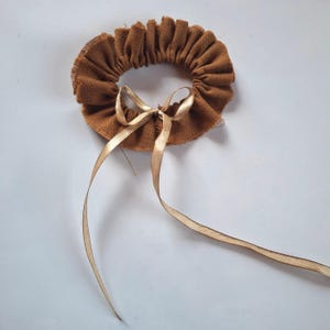 Könnte beinhalten: Ein kreisförmiges braunes Stoff-Haargummi mit gerüschtem, strukturiertem Rand und einem dünnen, glatten goldenen Band, das vorne zu einer Schleife gebunden ist. Das Accessoire hat ein gerafftes Design mit sichtbaren ausgefransten Kanten.