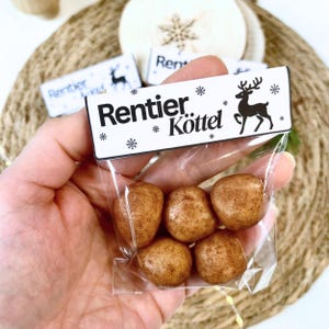 Könnte beinhalten: Ein klarer Plastikbeutel mit sechs braunen, abgerundeten Süßigkeiten. Der Beutel hat ein weißes Etikett mit dem Text "Rentier Köttel" und einer Rentiersilhouette. Die Süßigkeiten werden in einer Hand gehalten, mit anderen dekorativen Elementen im Hintergrund.