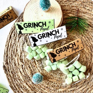 May include: A festive arrangement featuring green marshmallows in clear bags, accompanied by decorative signs that read "GRINCH Popel" with a Grinch silhouette and snowflakes. The signs are in green, white, and brown.