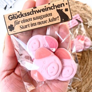 Könnte beinhalten: Eine transparente Plastiktüte mit drei rosa Schweinchen-Bonbons mit dunkleren rosa Akzenten. Ein kleines Holzschild mit deutschem Text und einer Schweinesilhouette ist an der Tüte befestigt.