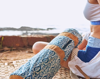May include: A blue and white patterned yoga mat bag with a tan leather accent and a zipper closure. The bag is lying on a woven mat. A white strap and a person wearing athletic clothing are also visible.