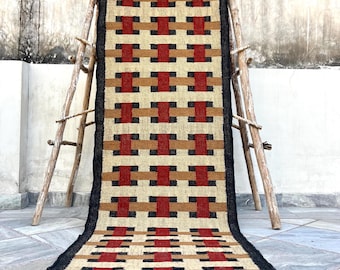 May include: A rectangular rug with a geometric pattern in red, black, tan, and brown. The rug is displayed on a wooden ladder. The rug has a black border and fringe.