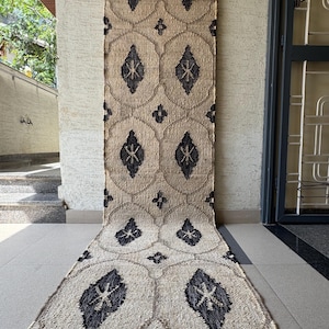 Op de afbeelding: Een beige en zwarte jute loper met een herhalend geometrisch patroon. Het ontwerp bevat donkere vormen in cirkelvormige en ruitvormige vormen. De loper hangt aan een houten roede tegen een getextureerde muur.
