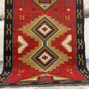 Puede incluir: Una alfombra rectangular tejida con un diseño geométrico. La alfombra presenta un fondo rojo con un patrón de diamante central en beige, negro y verde. Los bordes negros con formas de X blancas repetidas enmarcan los lados.