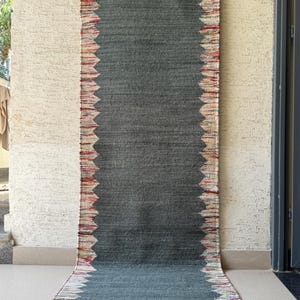 Puede incluir: Una alfombra larga y rectangular con un centro gris oscuro y un borde colorido con diseño en zigzag. La alfombra está hecha de tela tejida y se muestra verticalmente, mostrando su longitud y diseño. El borde presenta tonos rojos, blancos y amarillos.