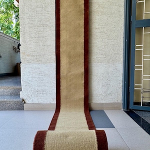 May include: A rolled-up beige and burgundy runner rug. The rug has a beige center with burgundy borders. The rug is hanging from a wooden rod and is displayed on a tiled floor. The rug is made of a woven material.