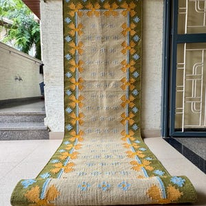 Puede incluir: Una alfombra de pasillo tejida en beige y verde con un estampado floral. La alfombra tiene un borde verde con diseños de flores amarillas y azules. La alfombra está enrollada en la parte inferior, mostrando el patrón y la textura.