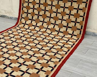 Könnte beinhalten: Ein rechteckiger Teppich mit geometrischem Muster. Der Teppich hat einen beigefarbenen Hintergrund mit braunen Quadraten und schwarzen Kreuzformen. Ein roter Rand umrahmt den Teppich, und die Kanten haben einen Fransen. Der Teppich besteht aus gewebtem Material.