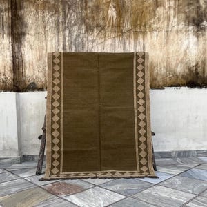 Op de afbeelding: Een rechthoekig bruin tapijt met een ruitpatroon in een lichtere bruintint. Het tapijt wordt gepresenteerd tegen een getextureerde muur en rust op een houten steun. Het tapijt heeft een franje langs de onderrand.