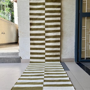 Op de afbeelding: Een lange, rechthoekige vloerkleed met afwisselende horizontale strepen in olijfgroen en wit. Het vloerkleed hangt verticaal, waarbij het onderste deel zich uitstrekt over een betegeld oppervlak. Het ontwerp van het vloerkleed is symmetrisch.