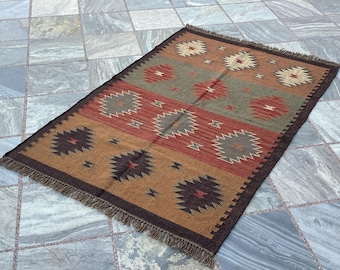 May include: Rectangular woven rug with a geometric pattern in shades of brown, red, and gray. The rug features a fringed edge and a repeating design of starburst and diamond shapes. The rug is placed on a tiled surface.