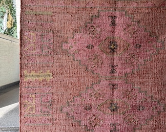 Puede incluir: Una alfombra tejida rectangular en tonos rosa empolvado y rosa, con un patrón geométrico repetitivo de diamantes en verde oliva apagado y marrón. El tejido tiene una superficie texturizada y gruesa con un tejido visible.