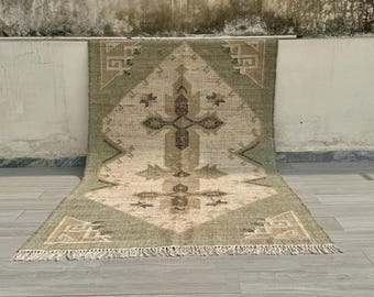May include: A rectangular woven rug featuring a sage green border and a central cream-colored diamond motif. The design includes dark brown geometric cross-shaped patterns and small star-like accents. The rug is finished with short fringe on both ends.