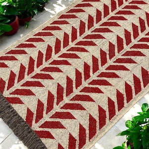 Puede incluir: Alfombra rectangular con fondo beige y un patrón repetido de formas rojas en forma de hojas. La alfombra tiene un borde con flecos en un tono marrón más oscuro. La alfombra está hecha de un material tejido y es adecuada para uso en interiores.