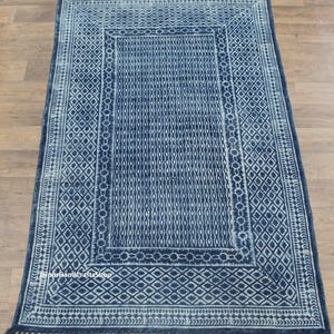 May include: A rectangular indigo blue rug with a geometric pattern. The rug features a central field of vertical lines, framed by diamond and floral borders. The rug has fringe on the short ends.