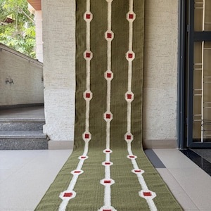 May include: A long, rectangular olive green runner rug featuring three vertical white stripes. Each white stripe is punctuated by a repeating pattern of small, solid red squares enclosed within white circular outlines, creating a geometric design.