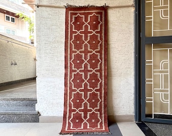 May include: A rectangular runner rug featuring a deep terracotta red base with a repeating white geometric star and cross pattern. The rug is finished with a thin white border and short fringe along the top and bottom edges.