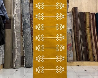 May include: Mustard yellow rectangular rug with white geometric patterns. The rug is held up in front of a shelf with rolled-up rugs. The white patterns are symmetrical and repeated across the length of the rug. The rug has a fringe at the bottom.