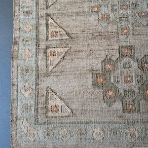 May include: A section of a woven rug with a muted colour palette. The rug features a light grey background with geometric patterns in shades of brown, green, and blue. The border has a light blue hue with decorative elements. The rug has a fringe.