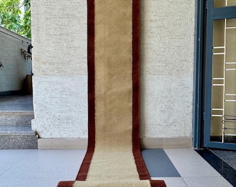 May include: A long, rectangular runner rug featuring a central tan panel flanked by two dark brown vertical borders. The rug is suspended from a wooden rod at the top and rolls onto the floor at the bottom, displaying a woven texture.