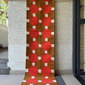 May include: A rectangular, woven wall hanging with a geometric pattern. The design features alternating brown and red diamond shapes, separated by white squares. The rug is hanging on a wooden rod against a textured wall.
